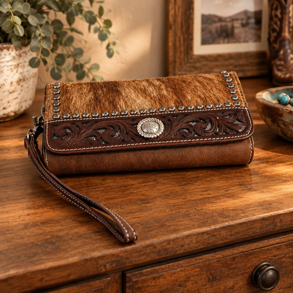 A brown leather wristlet clutch with a cowhide-look top panel, tooled leather detailing, a silver-tone concho.