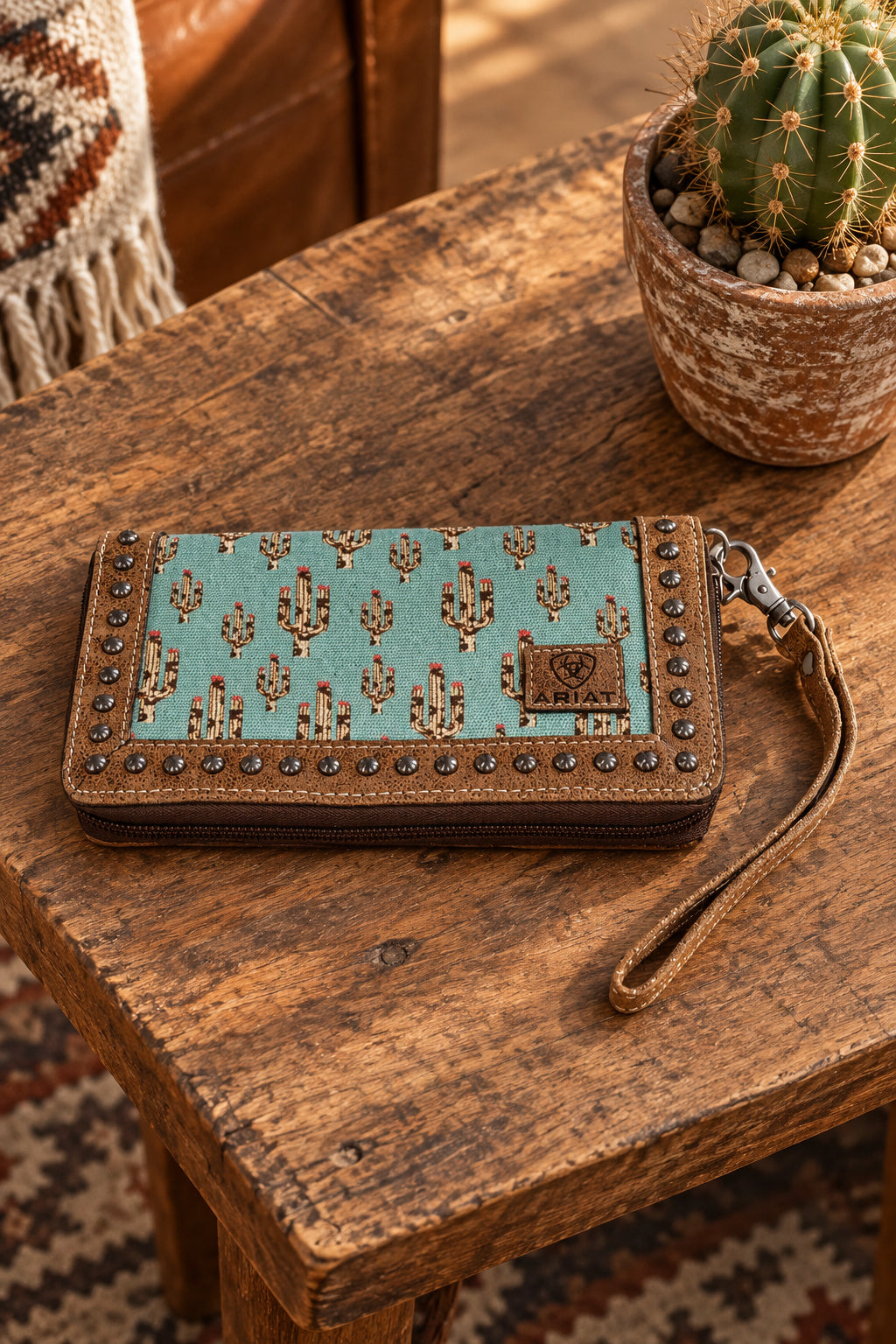 A western-style clutch wallet with a turquoise and brown cactus pattern, studded leather trim, and a wrist strap.