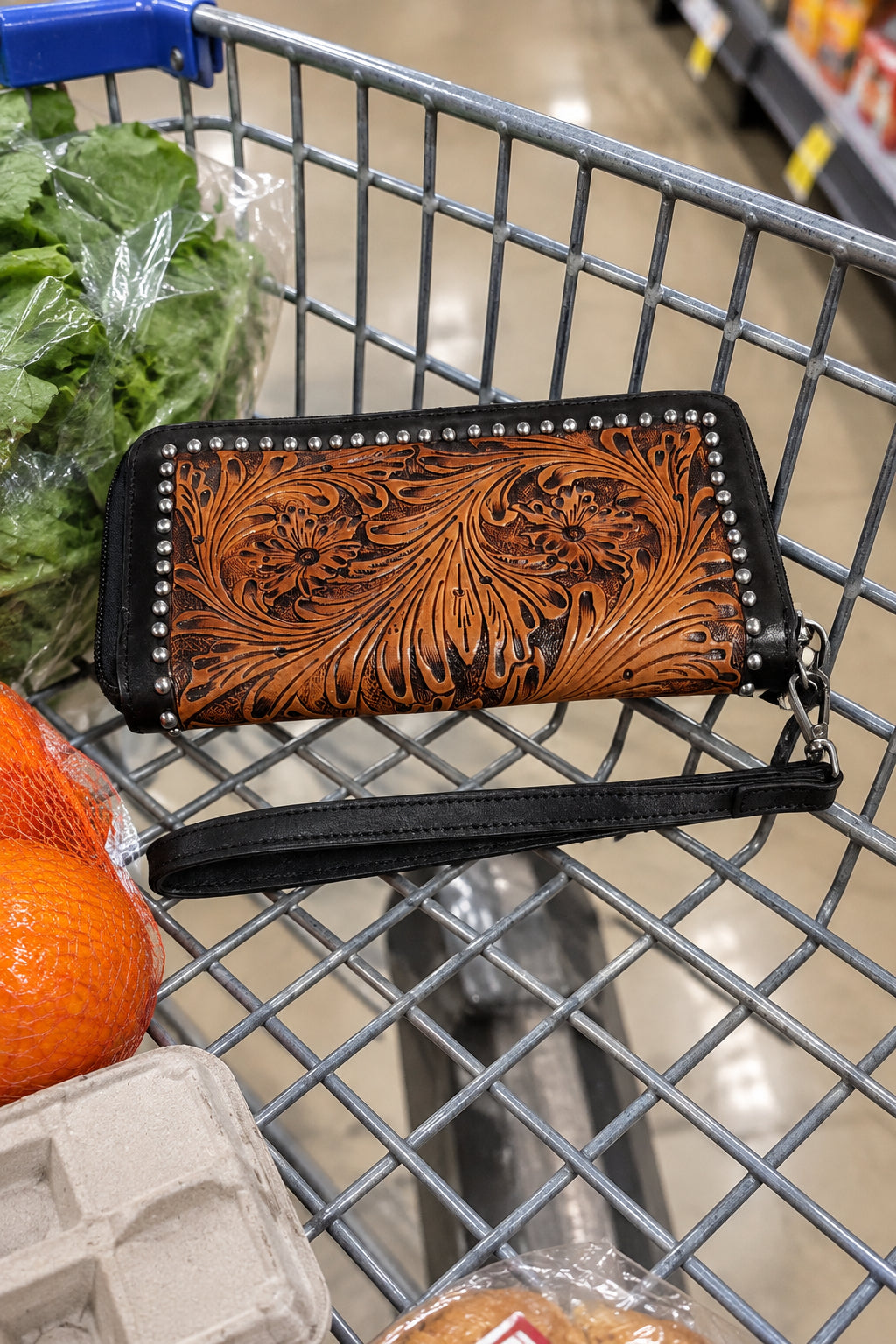 A brown tooled leather zip-around wallet with floral and scroll engravings, black leather trim, silver-tone studs, and a matching black wristlet strap.