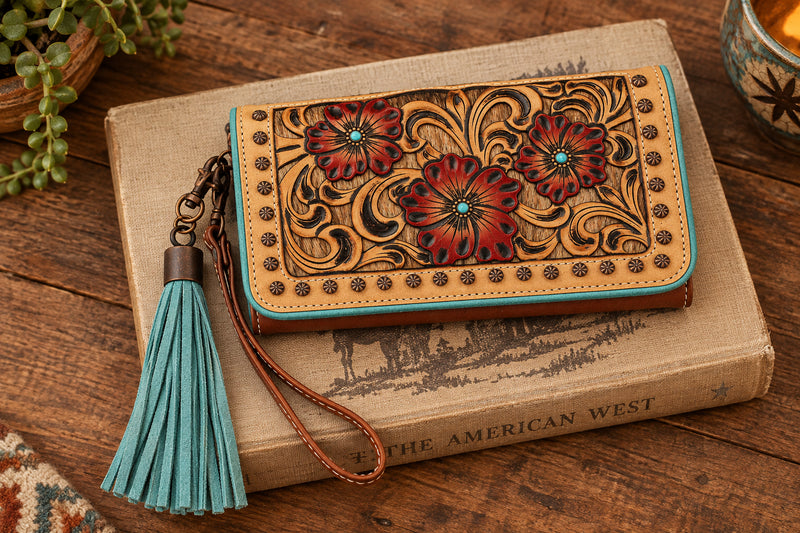 A tan calf hair clutch wallet with tooled floral overlay featuring red flowers and a turquoise fringe tassel, with a brown faux leather Snap closure.